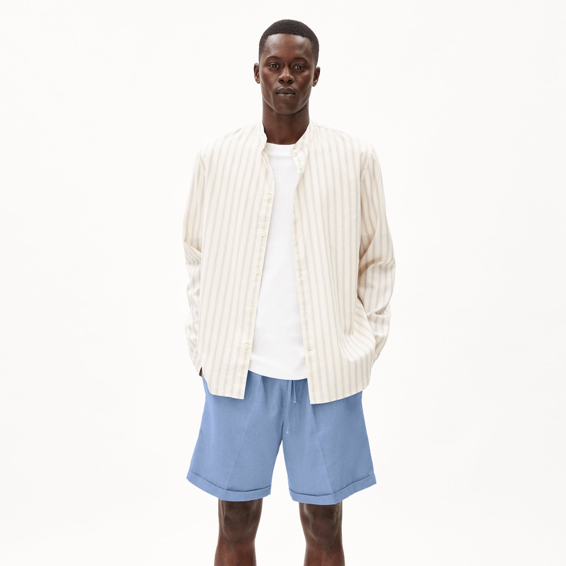 Ein Mann trägt ein gestreiftes Hemd, ein weißes T-Shirt und blaue Shorts vor einem weißen Hintergrund.