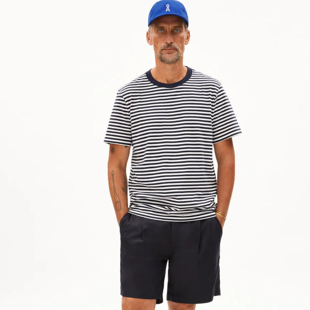 Ein Mann steht vor einem weißen Hintergrund und trägt eine blaue Mütze, ein marineblau-weiß gestreiftes T-Shirt, marineblaue Shorts, leuchtend orangefarbene Socken und lila Slipper. Seine Hände stecken in den Taschen, und er hat sichtbare Armtattoos.