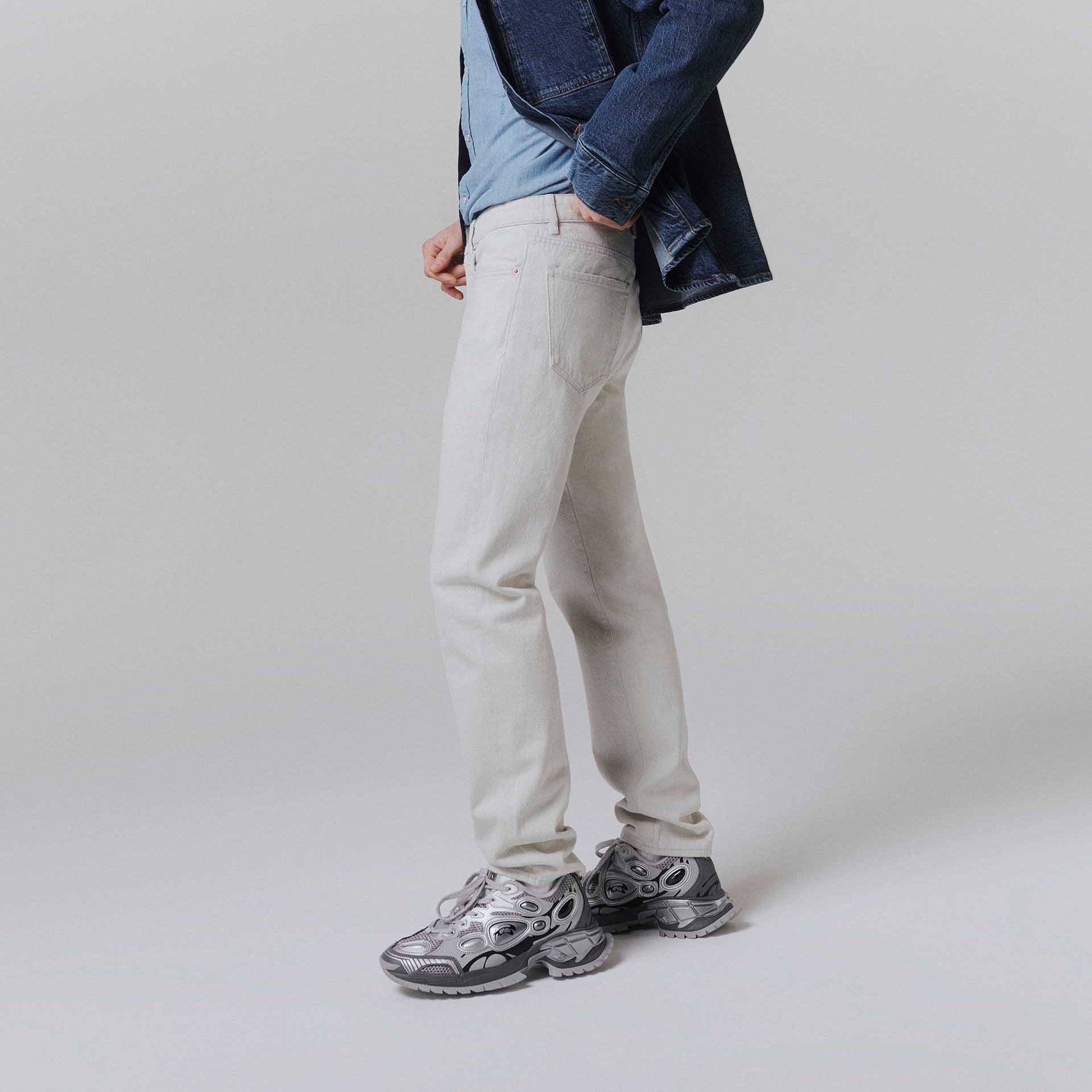 Person mit weißer Jeans, Jeansjacke und silbernen Turnschuhen steht seitlich vor neutralem Hintergrund.