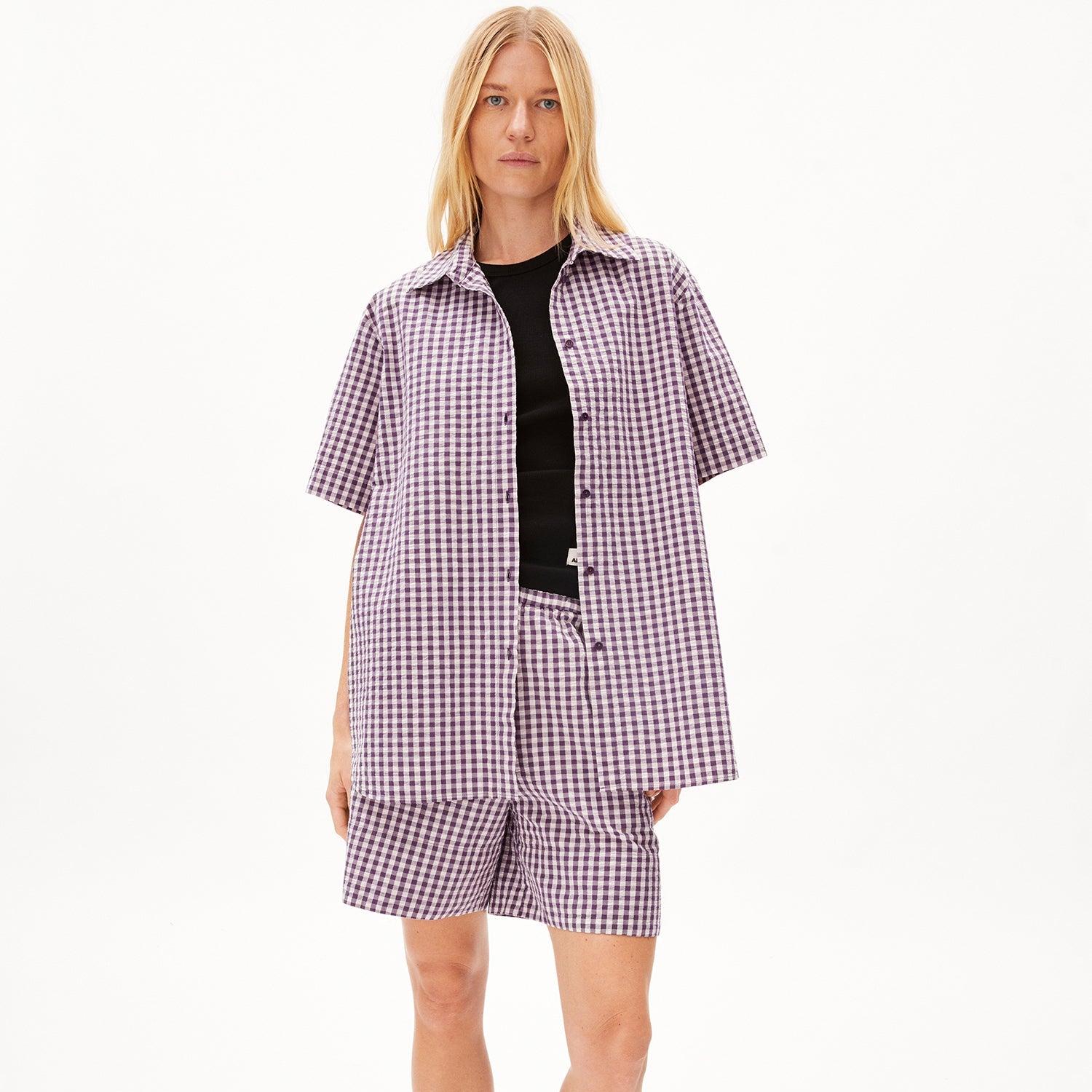 Frau in lila kariertem Hemd und Shorts, mit schwarzen Top darunter, vor weißem Hintergrund.