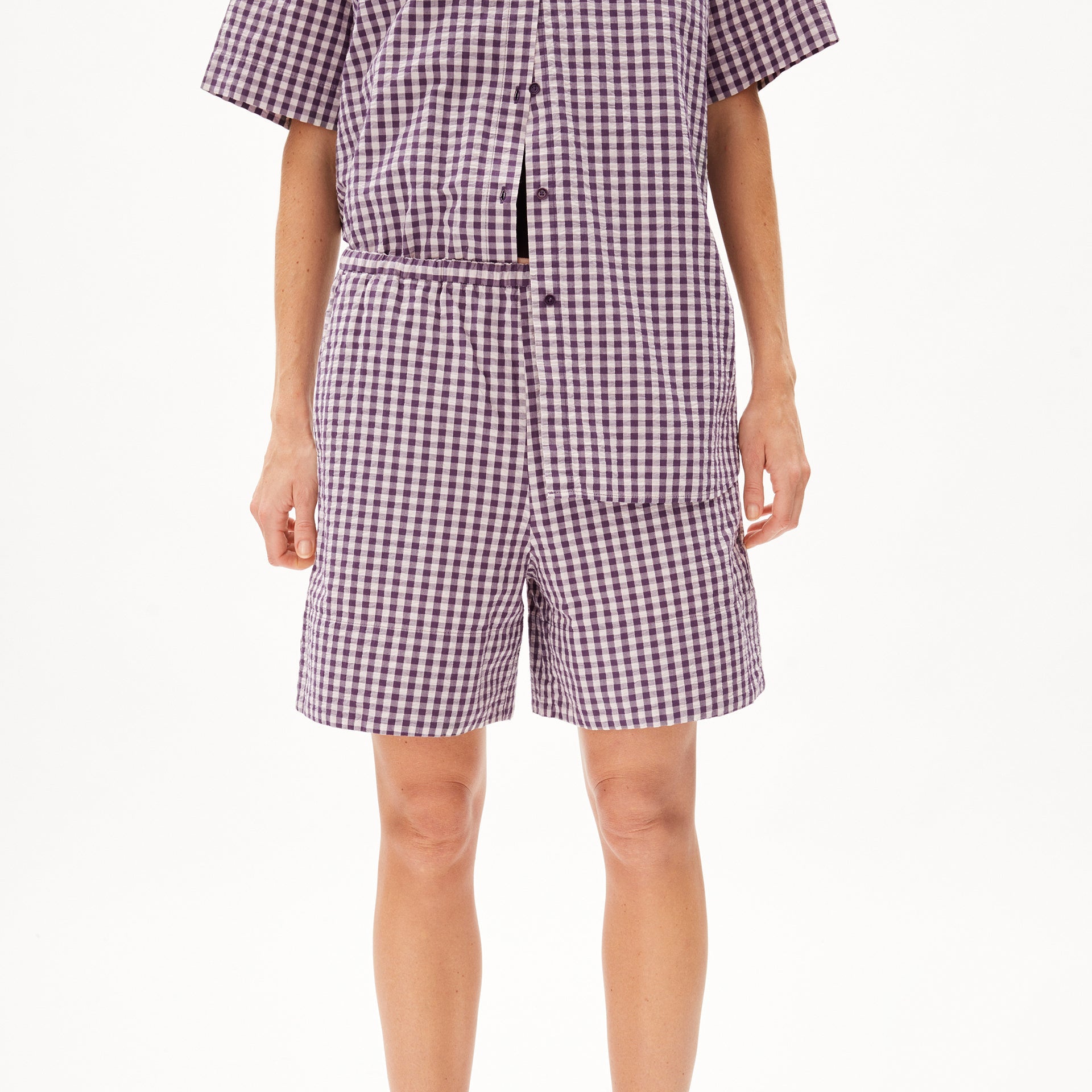 Person in einem lila-weiß karierten Outfit mit kurzen Ärmeln und Shorts, stehend vor weißem Hintergrund.