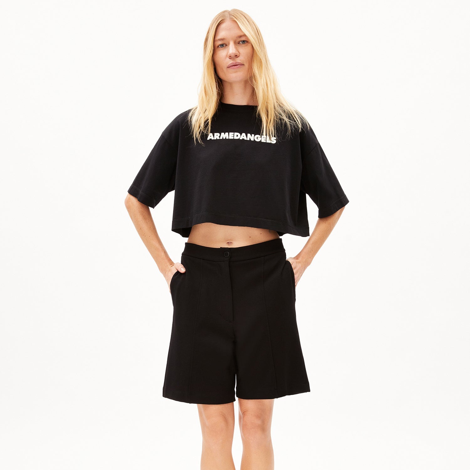 Person in schwarzem Crop-Top mit "ARMEDANGELS"-Schriftzug und schwarzer Shorts, steht mit Händen in den Taschen.