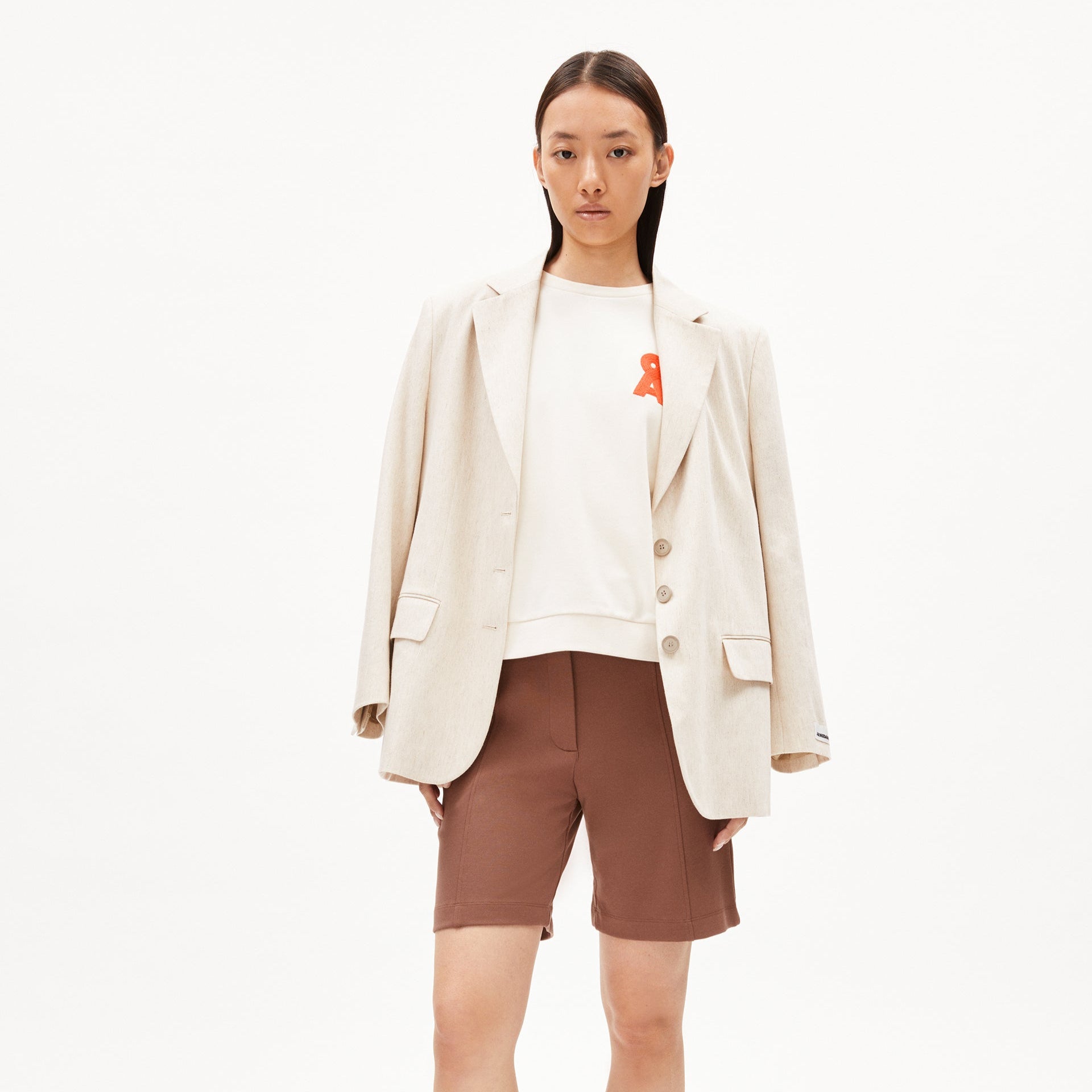 Frau in beigem Blazer, weißem Shirt mit rotem Detail und braunen Shorts steht vor weißem Hintergrund.