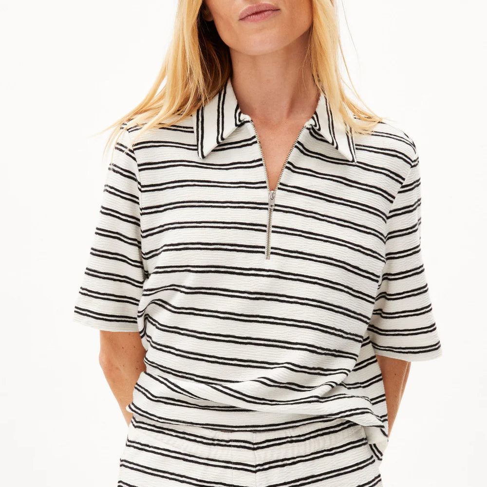 Eine Frau mit langen blonden Haaren trägt ein kurzärmeliges, weißes Hemd mit schwarzen Querstreifen und einem Kragen mit Reißverschluss, gepaart mit einer passenden gestreiften Hose, vor einem schlichten weißen Hintergrund.