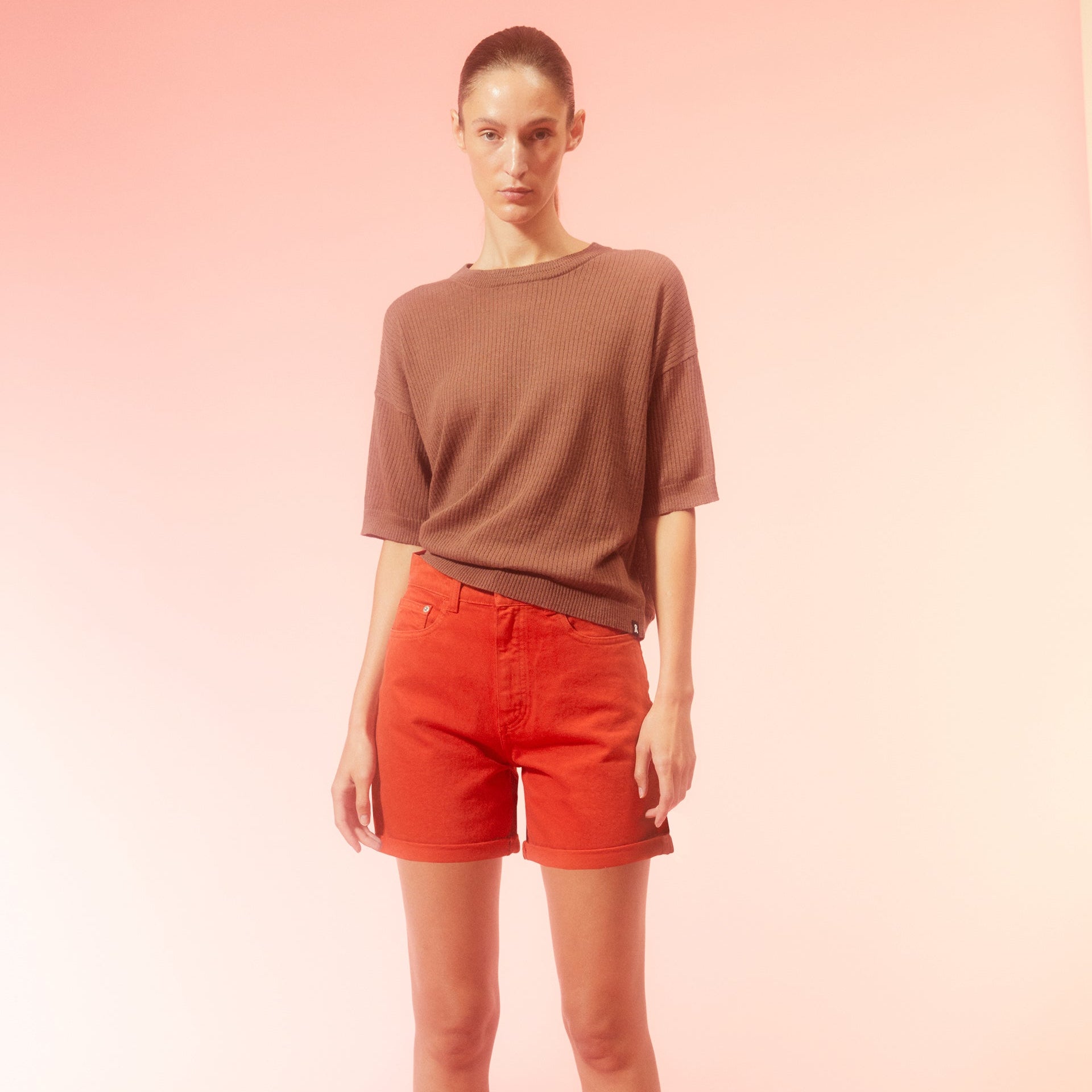 Frau in braunem Shirt und roten Shorts steht vor einem rosa Hintergrund.