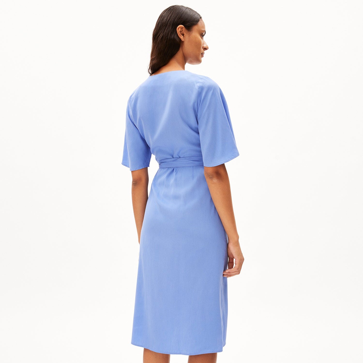 Frau in einem hellblauen Kleid mit Gürtel, von hinten fotografiert, vor weißem Hintergrund.