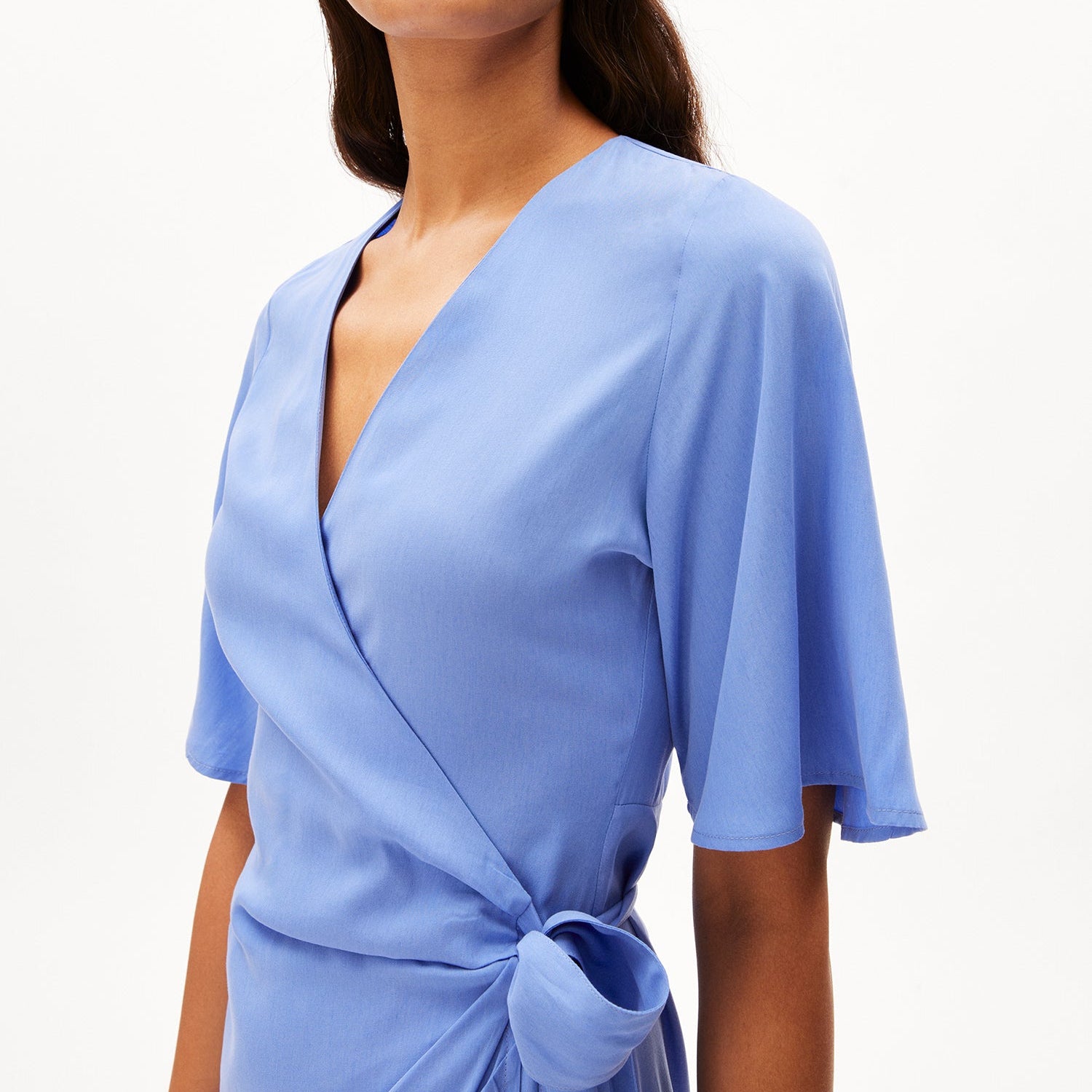 Frau in einem blauen Wickelkleid mit V-Ausschnitt und kurzen Ärmeln vor einem weißen Hintergrund.