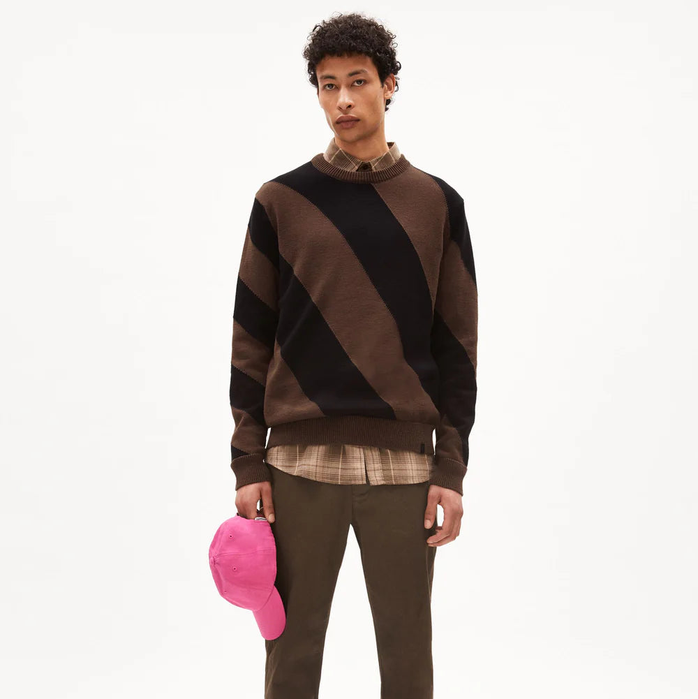 Eine junge Person steht mit dem Gesicht nach vorne, trägt einen übergroßen braun-schwarz diagonal gestreiften Pullover über einem karierten Hemd, eine olivgrüne Hose, graue Turnschuhe und hält eine leuchtend pinke Baseballmütze in der Hand.