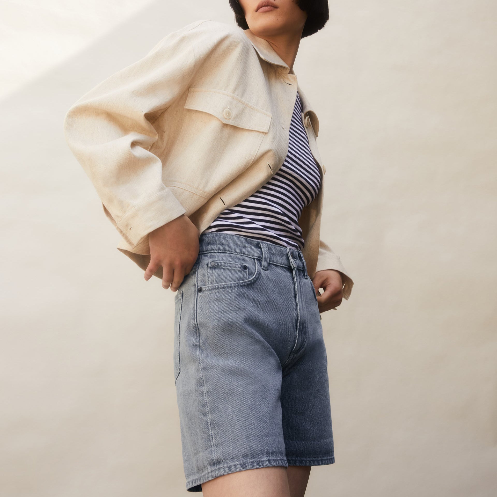 Person in beige Jacke und gestreiftem Top, trägt graue Jeansshorts vor neutralem Hintergrund.