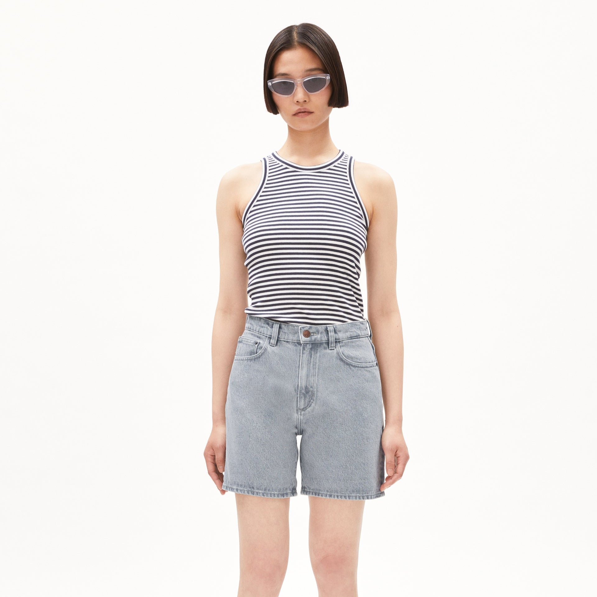 Frau in gestreiftem ärmellosem Oberteil und Jeansshorts trägt Sonnenbrille vor weißem Hintergrund.