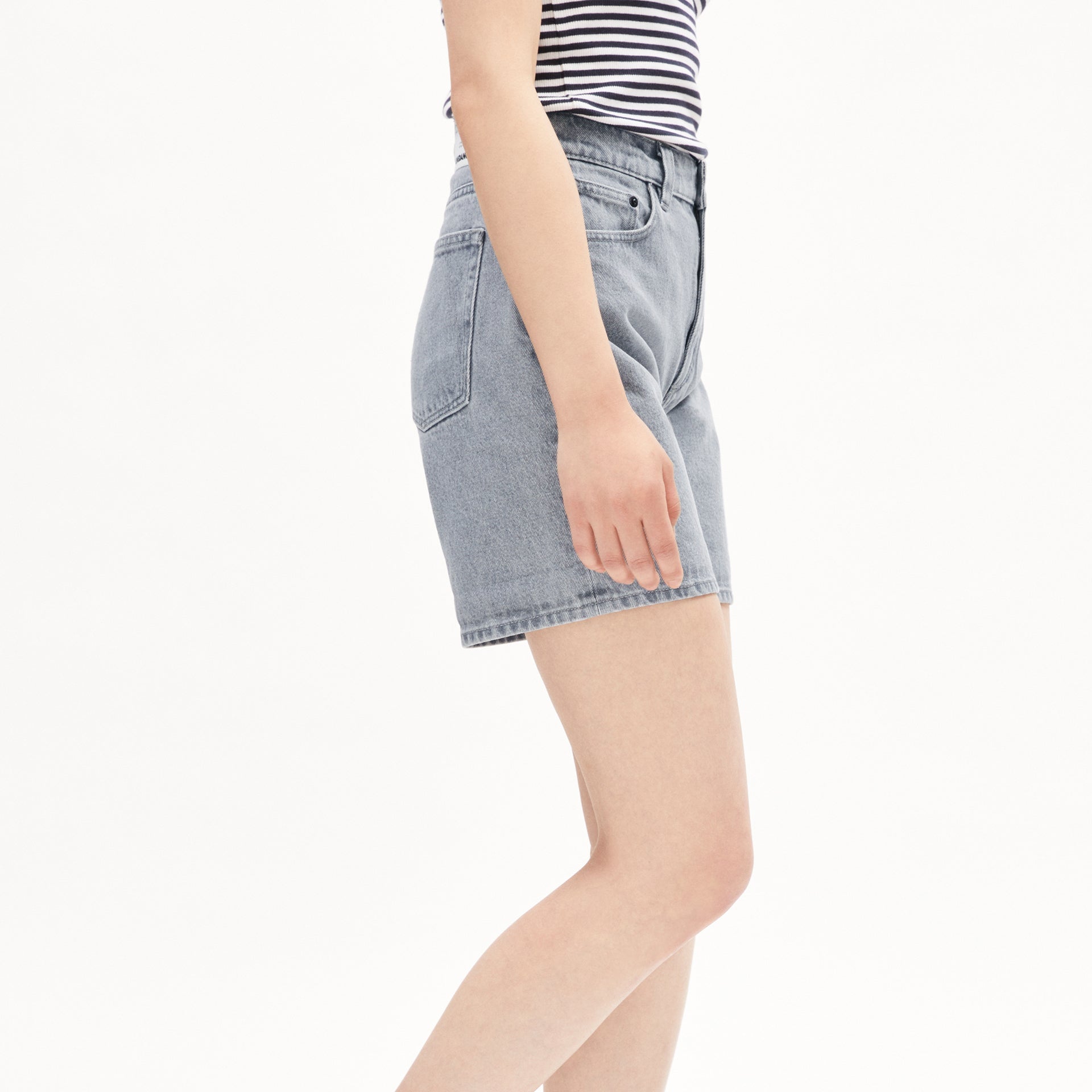 Person in gestreiftem Shirt und blauer Jeansshorts steht seitlich zu sehen vor weißem Hintergrund.