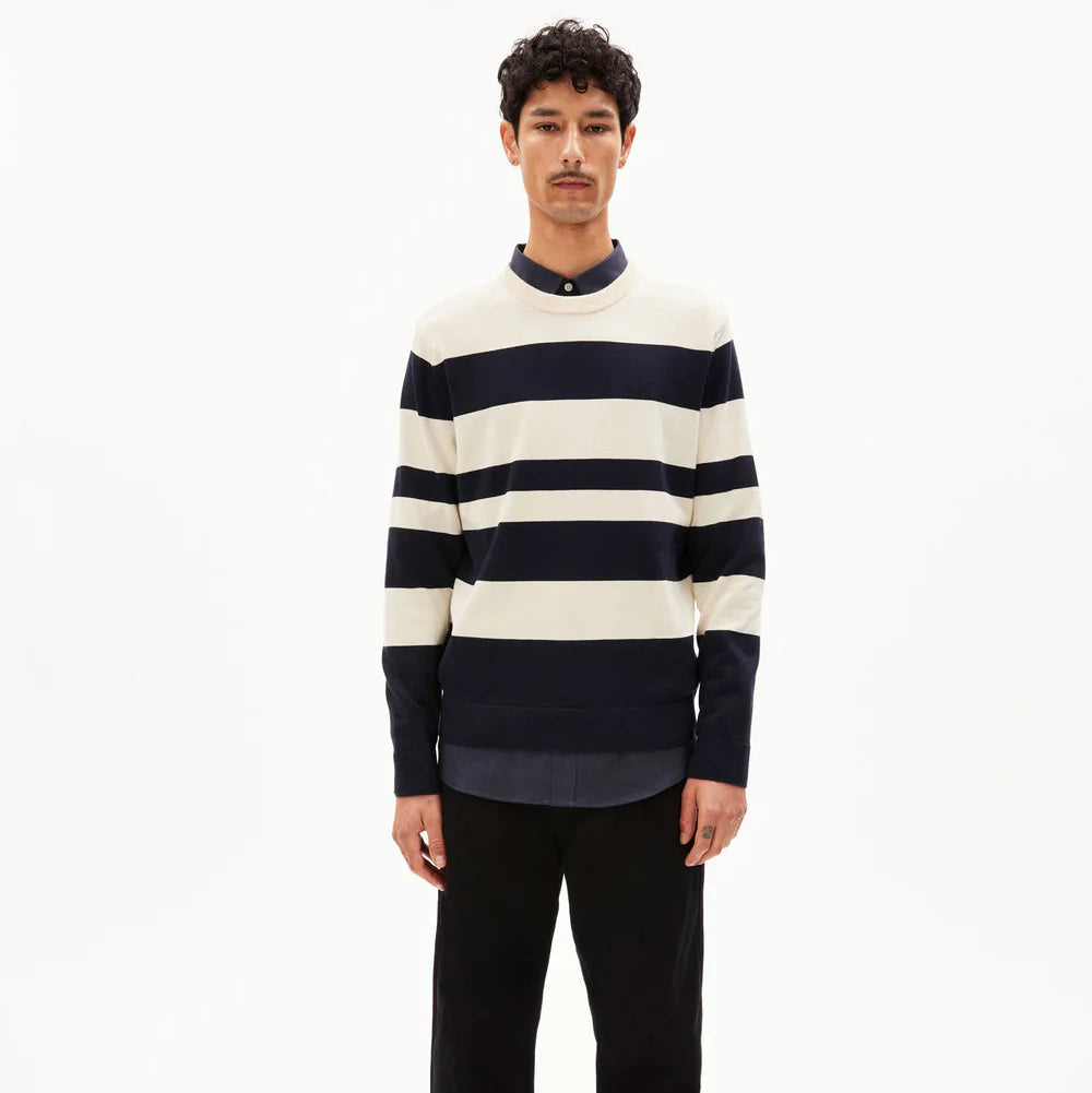 Ein Mann steht mit dem Gesicht nach vorne, trägt ein langärmeliges Hemd mit breiten schwarzen und weißen Querstreifen, eine schwarze Hose und silberne Turnschuhe vor einem schlichten weißen Hintergrund.