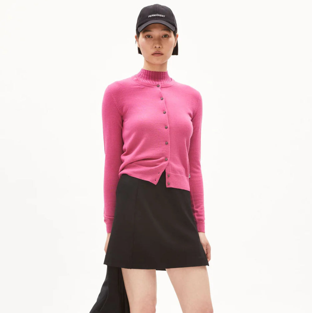 Eine Person, die eine rosa Strickjacke mit Knöpfen, einen schwarzen Minirock, schwarz-weiße Halbschuhe mit schwarzen Socken und eine schwarze Mütze trägt, steht vor einem schlichten weißen Hintergrund und hält ein schwarzes Kleidungsstück in einer Hand.