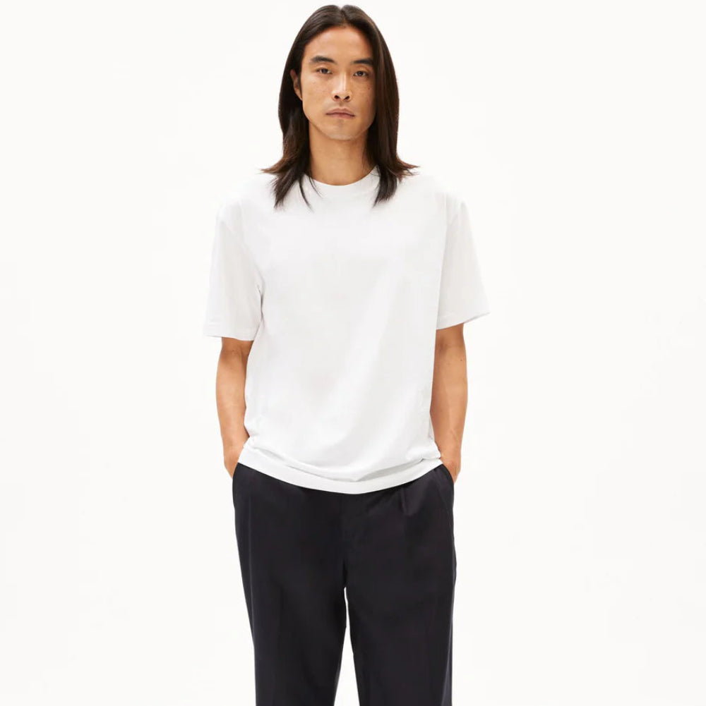 Eine Person mit langen dunklen Haaren, die ein schlichtes weißes T-Shirt, eine weite schwarze Hose und graue Turnschuhe trägt, steht mit dem Gesicht nach vorne und den Händen in den Taschen vor einem schlichten weißen Hintergrund.