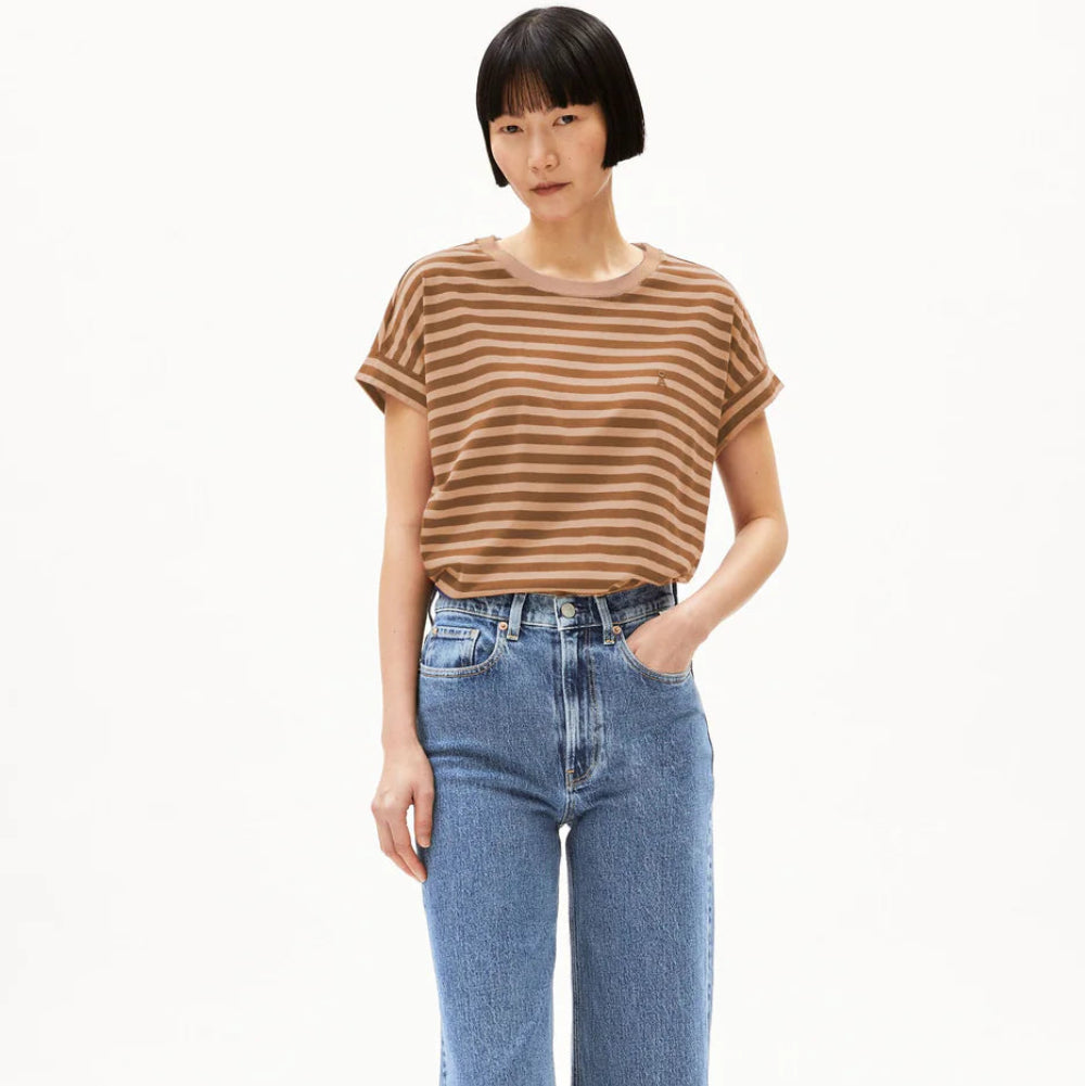 Eine Person mit kurzen schwarzen Haaren trägt ein braun-beige gestreiftes T-Shirt, hochgeschnittene blaue Jeans mit weitem Bein und schwarze Slide-Sandalen und steht vor einem schlichten weißen Hintergrund.