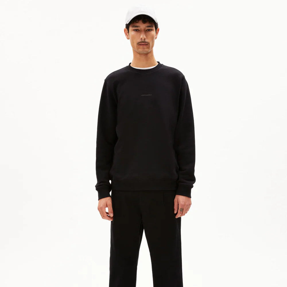 Ein Mann steht mit dem Gesicht nach vorne, trägt ein schwarzes Sweatshirt, eine schwarze Hose, schwarze Schuhe und eine hellgraue Baseballmütze vor einem schlichten weißen Hintergrund.