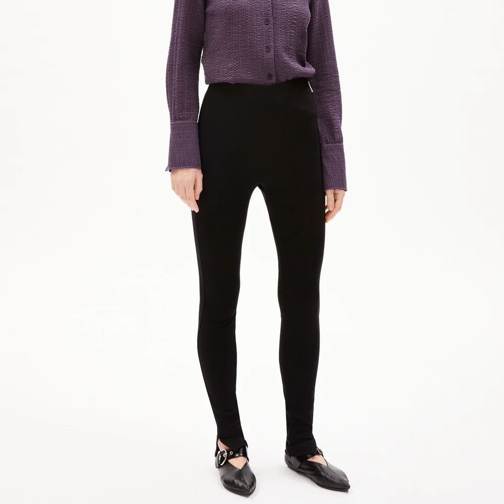 Eine Frau steht vor einem schlichten weißen Hintergrund und trägt ein lilafarbenes, strukturiertes Hemd zum Aufknöpfen, eine schwarze, eng anliegende Hose und schwarze Schuhe. Sie hat glattes, schulterlanges Haar und einen neutralen Gesichtsausdruck.