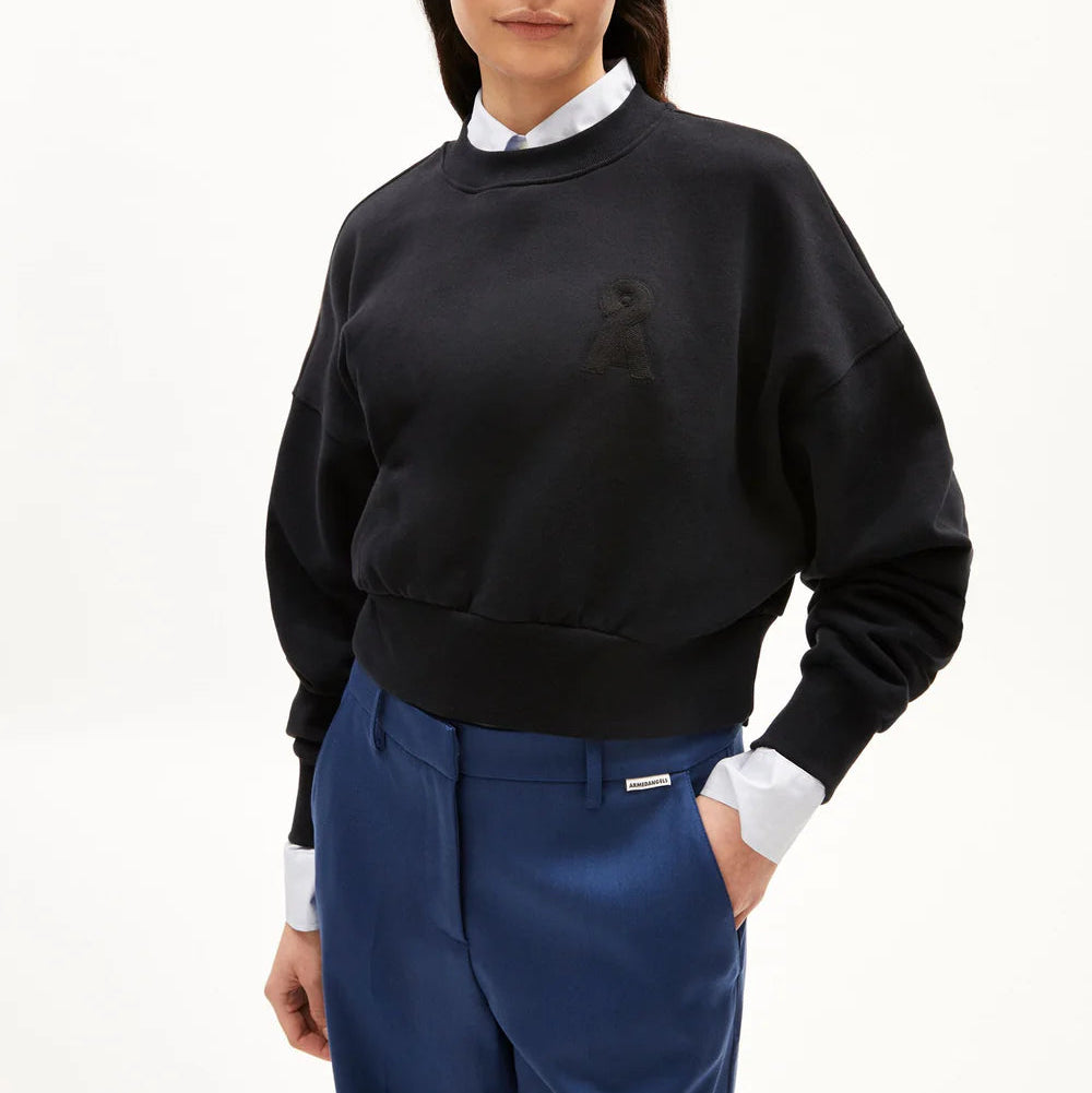Eine Frau trägt ein schwarzes, langärmeliges Sweatshirt mit einem dezent gestickten Logo und eine königsblaue Hose mit hoher Taille. Sie hat eine Hand in ihrer Tasche und steht vor einem schlichten weißen Hintergrund.