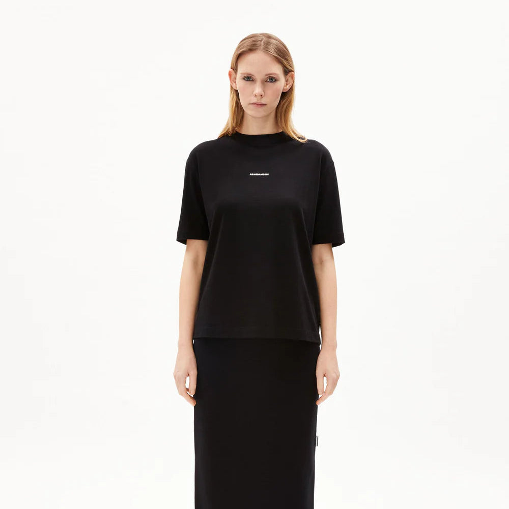 Eine Frau steht mit dem Gesicht nach vorne, trägt ein lockeres schwarzes T-Shirt, einen passenden schwarzen Midirock und schwarz-weiße Halbschuhe vor einem schlichten weißen Hintergrund.