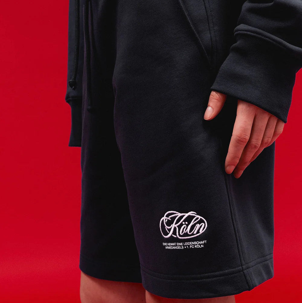 Person, die schwarze Shorts und ein schwarzes Sweatshirt trägt, vor einem leuchtend roten Hintergrund. Die Shorts sind mit weißem Text und einem stilisierten Logo in der Nähe des Saums am linken Bein versehen.