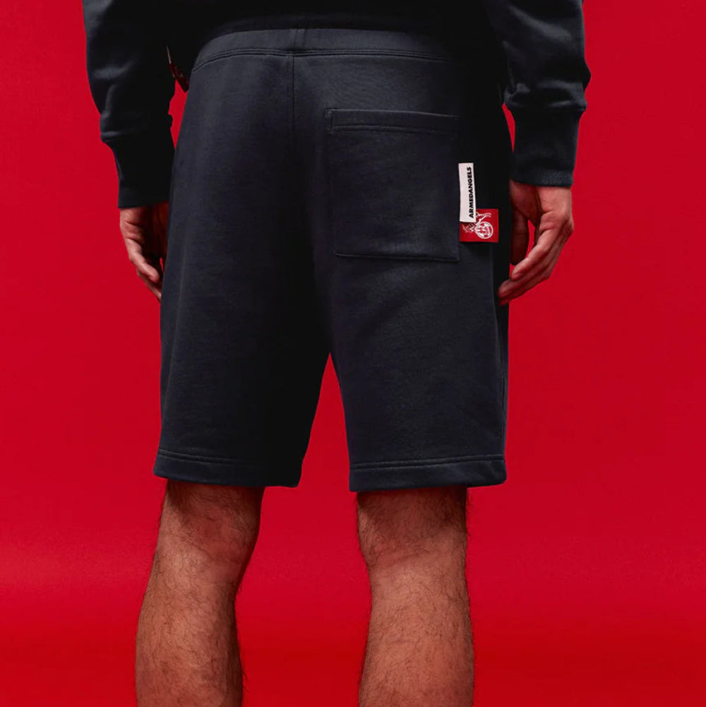 Eine Person steht vor einem roten Hintergrund und trägt von der Taille abwärts schwarze Shorts, schwarze Socken mit weißem Muster und schwarze Pantoffeln. An der rechten Tasche der Shorts ist ein Etikett zu sehen.