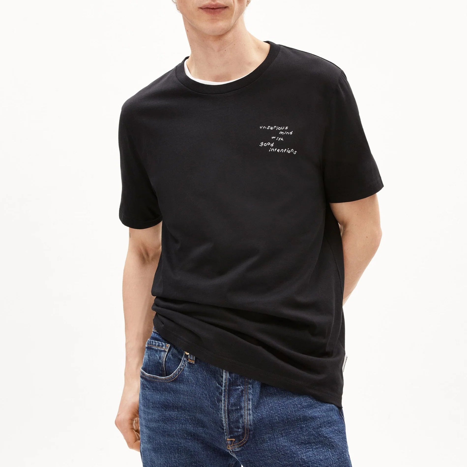 Ein Mann mit lockigem blondem Haar trägt ein schwarzes T-Shirt mit kleiner weißer Schrift auf der Brust und blaue Jeans, die vor einem schlichten weißen Hintergrund stehen.