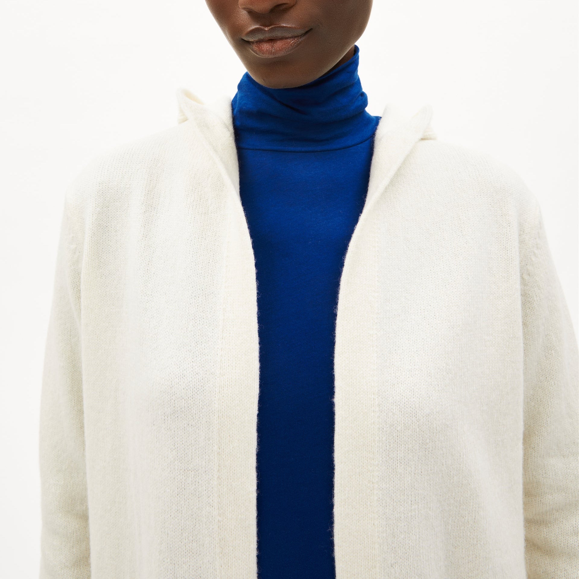 Person in cremefarbener Strickjacke mit blauem Rollkragenpullover darunter, Gesicht teilweise sichtbar.