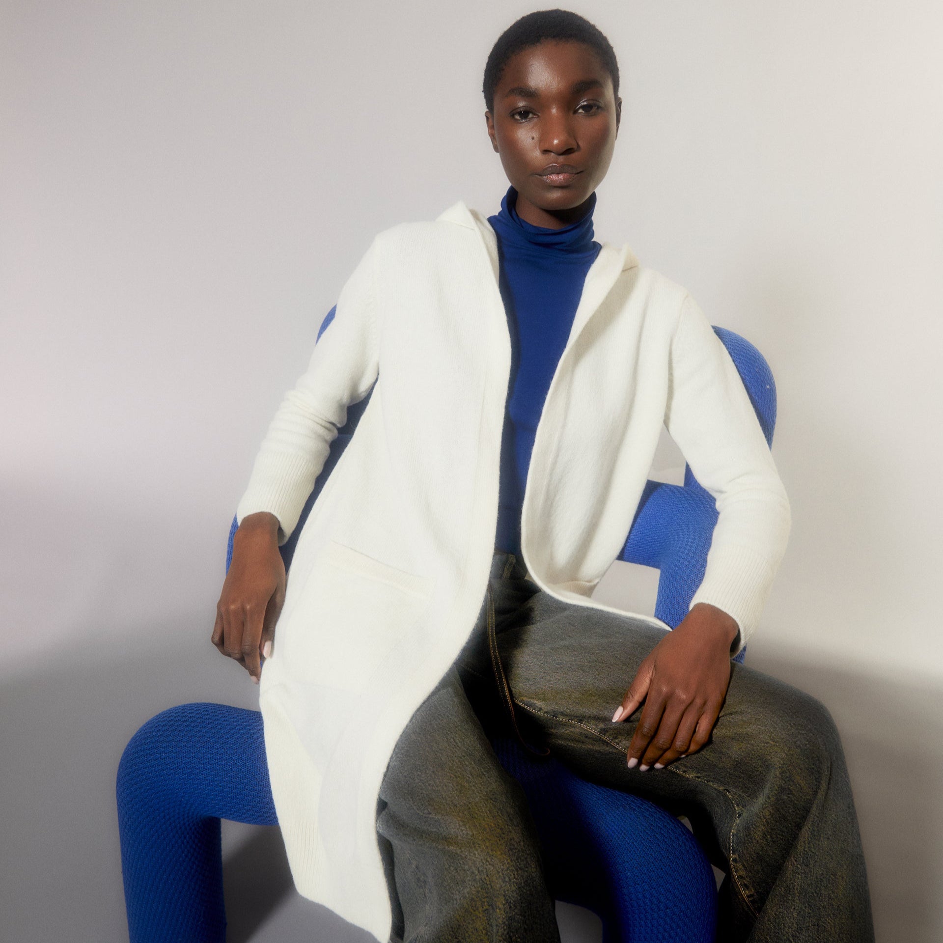 Person in weißem Mantel und blauem Rollkragenpullover sitzt auf einem blauen, modernen Stuhl.