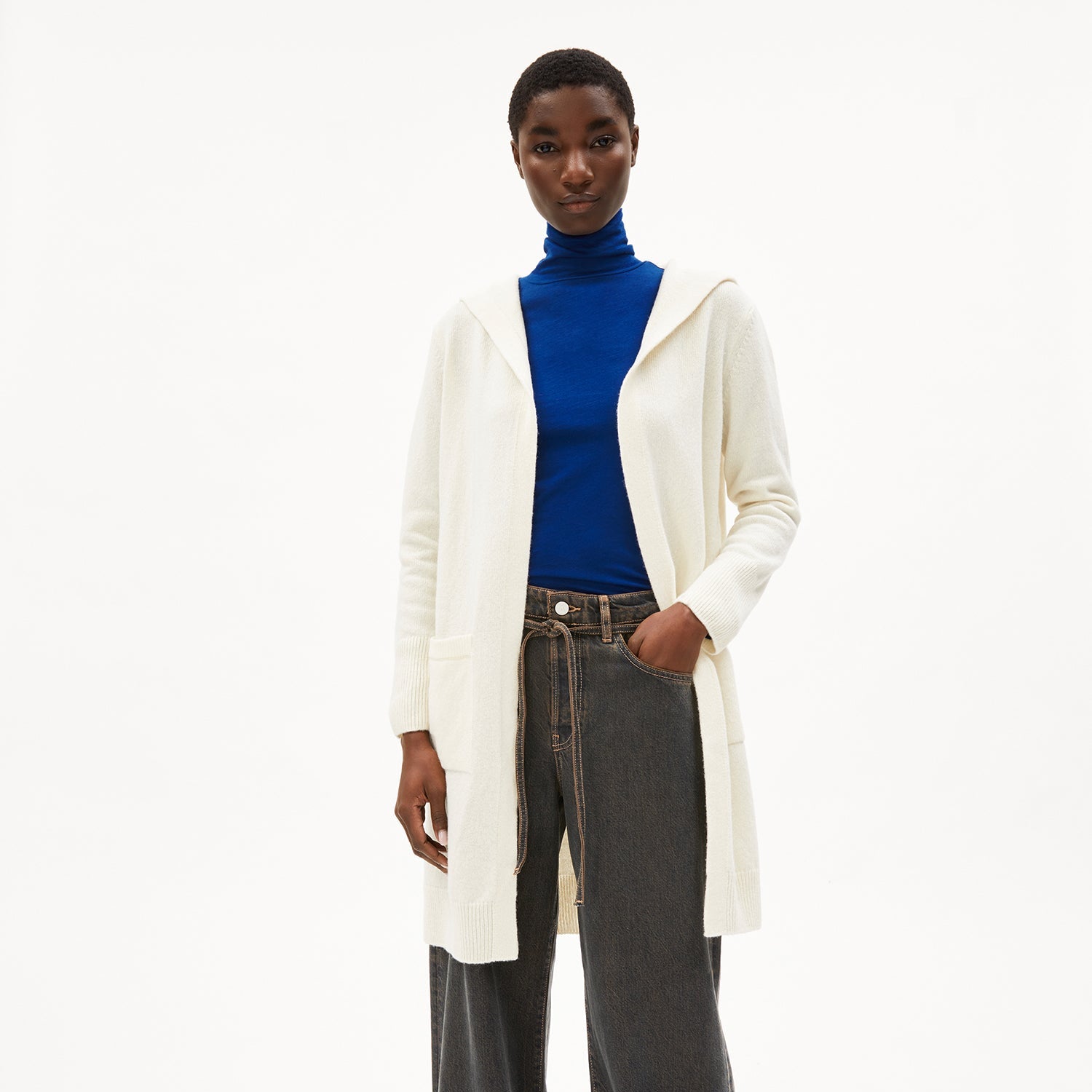Person in weißem Cardigan, blauem Rollkragenpullover und dunkler Jeans vor weißem Hintergrund.