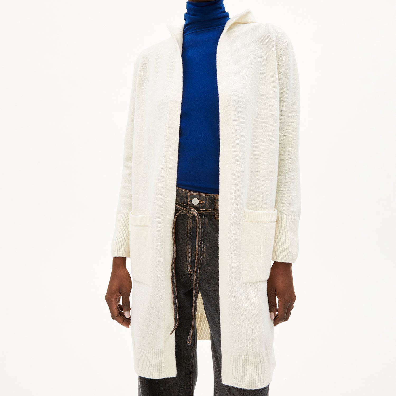 Person trägt einen weißen Strickmantel über einem blauen Rollkragenpullover und dunklen Jeans mit gebundenem Gürtel.