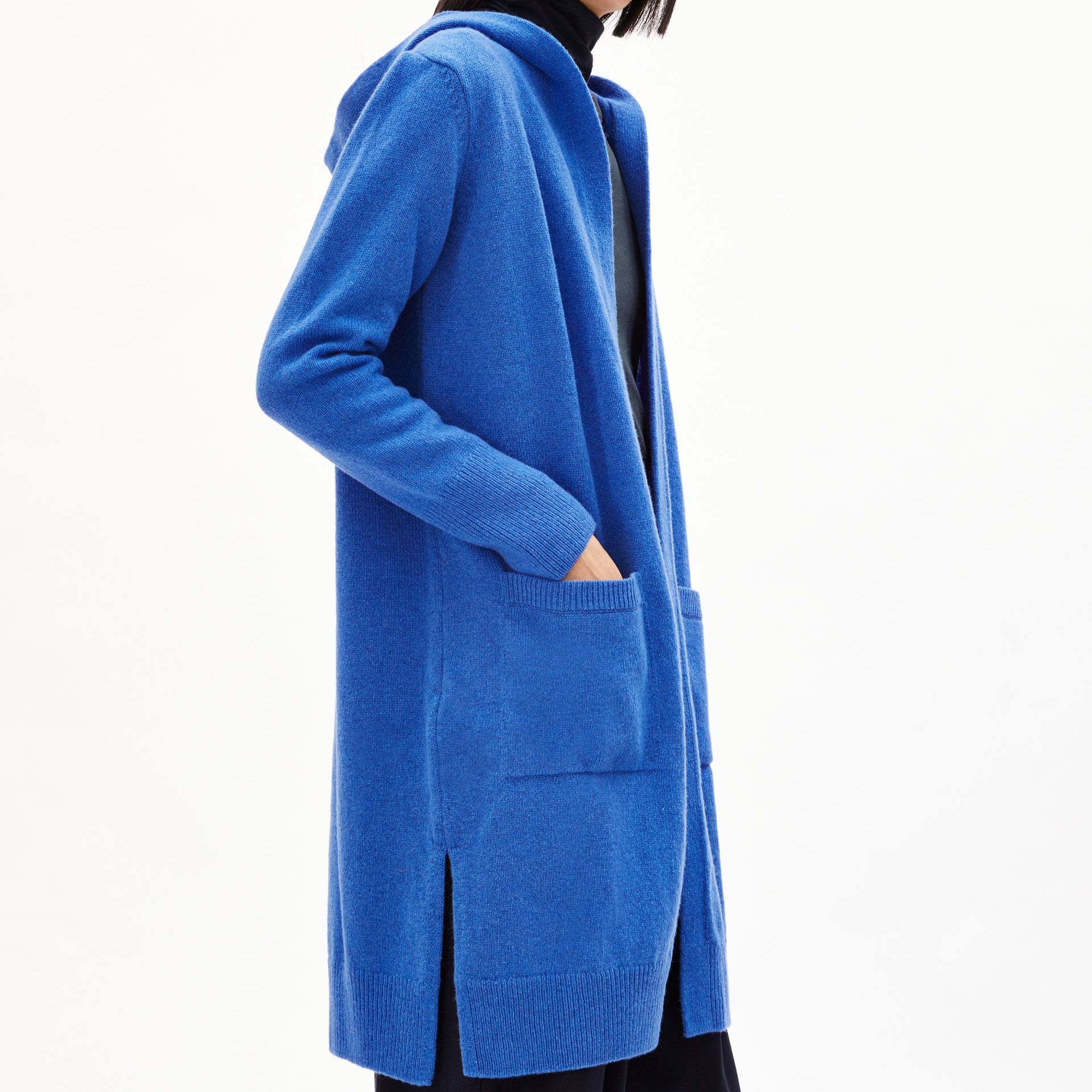 Eine Person trägt einen blauen langen Strickcardigan mit Kapuze und Taschen, die Hand in einer Tasche.