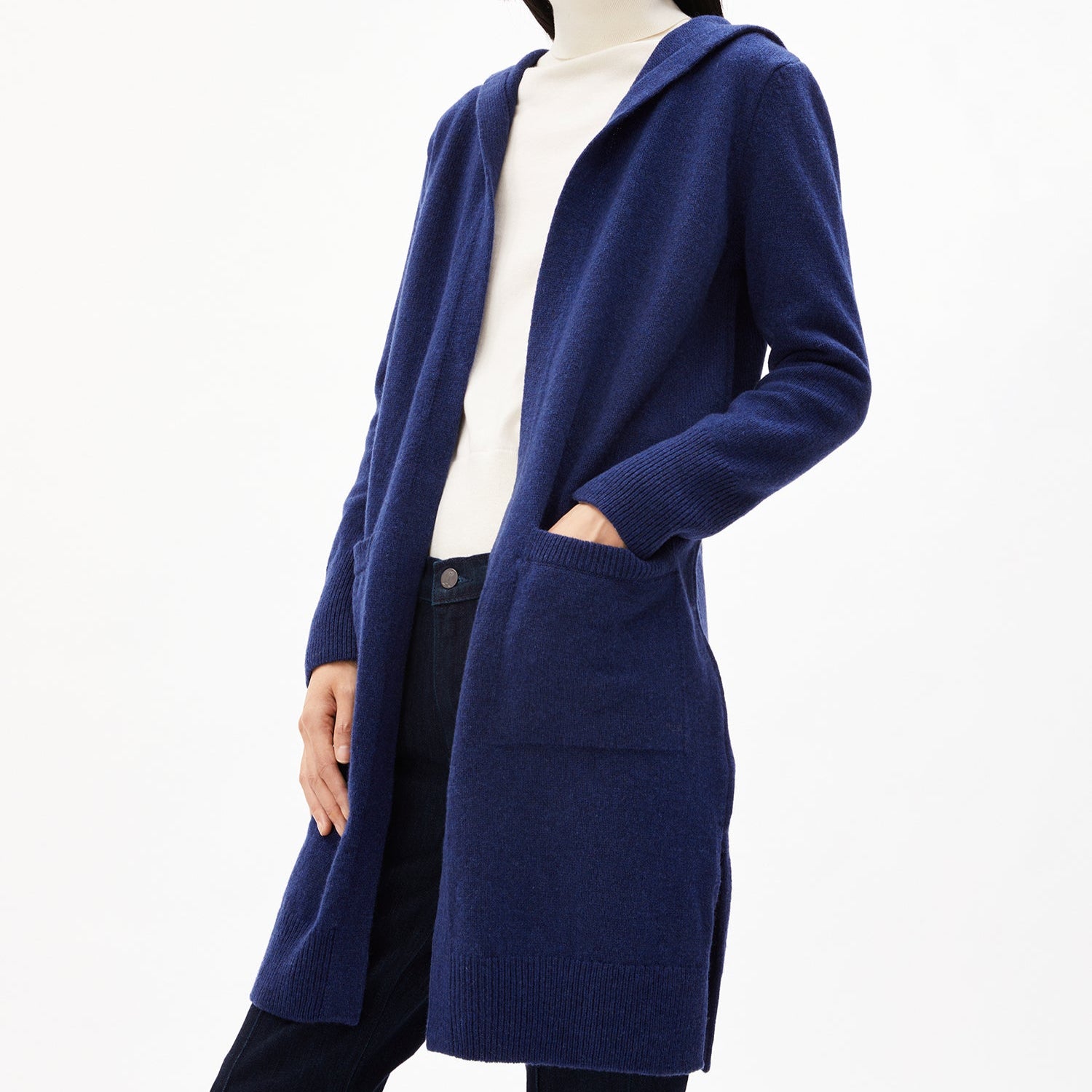 Person trägt einen langen blauen Cardigan aus Wolle über einem weißen Rollkragenpullover und schwarzen Jeans.