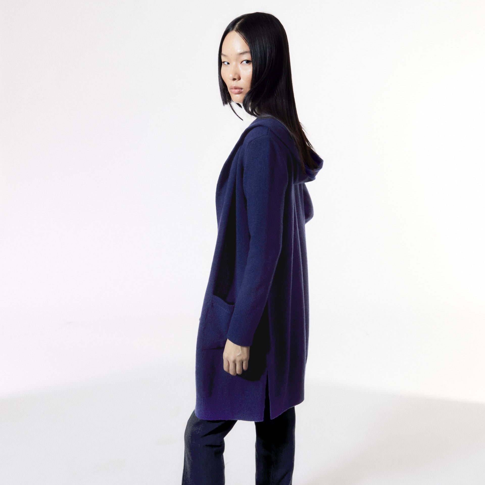 Eine Frau in einem langen, blauen Cardigan schaut über die Schulter in einem minimalistischen, weißen Hintergrund.
