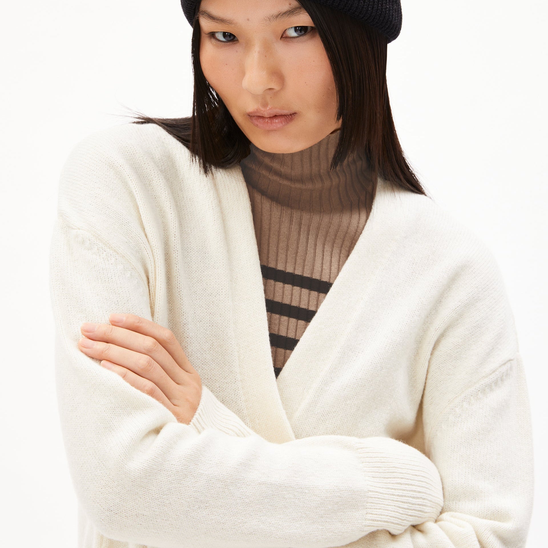 Person mit schwarzer Mütze und weißem Strickpullover, darunter ein gestreifter Rollkragenpullover, verschränkte Arme.