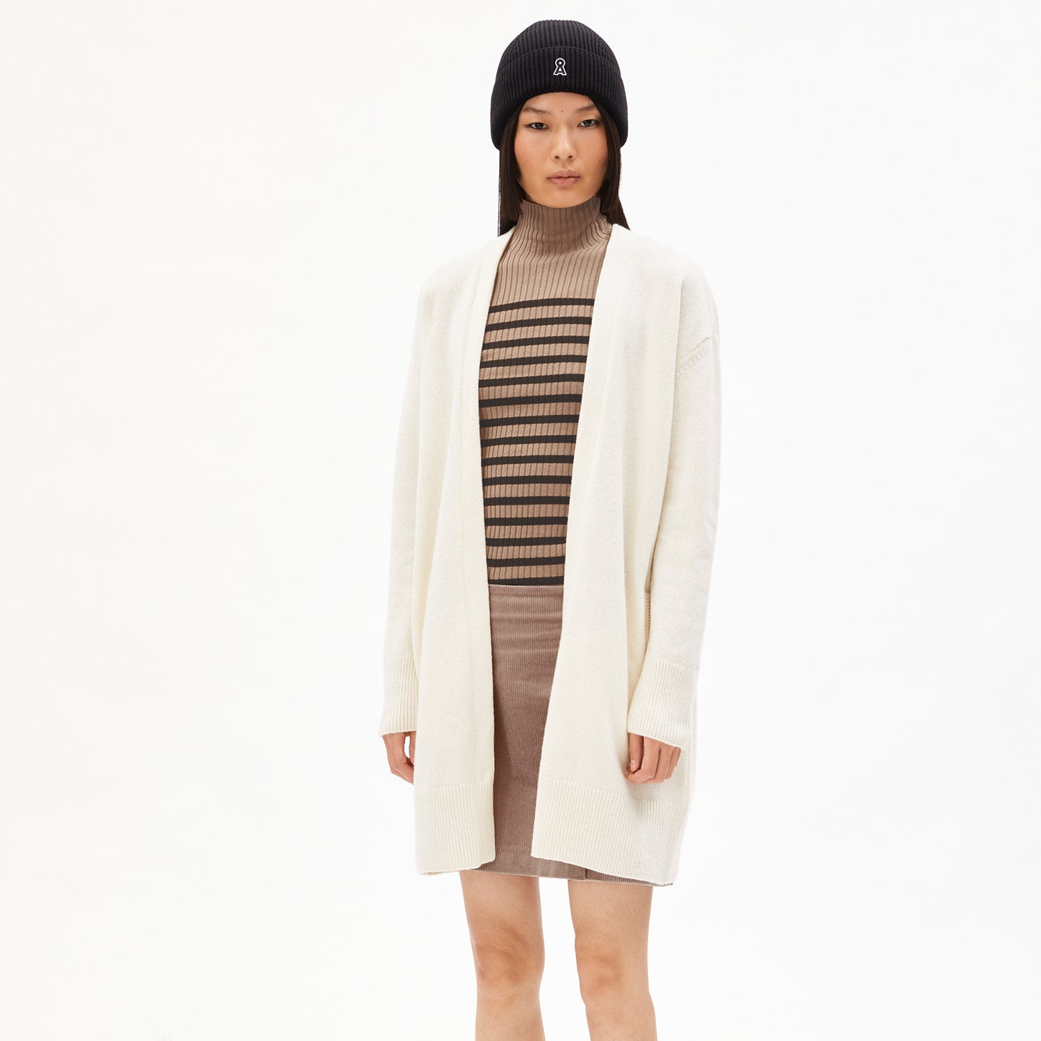 Frau trägt eine weiß-beige Strickjacke, gestreiftes Oberteil, Rock und schwarze Mütze vor weißem Hintergrund.