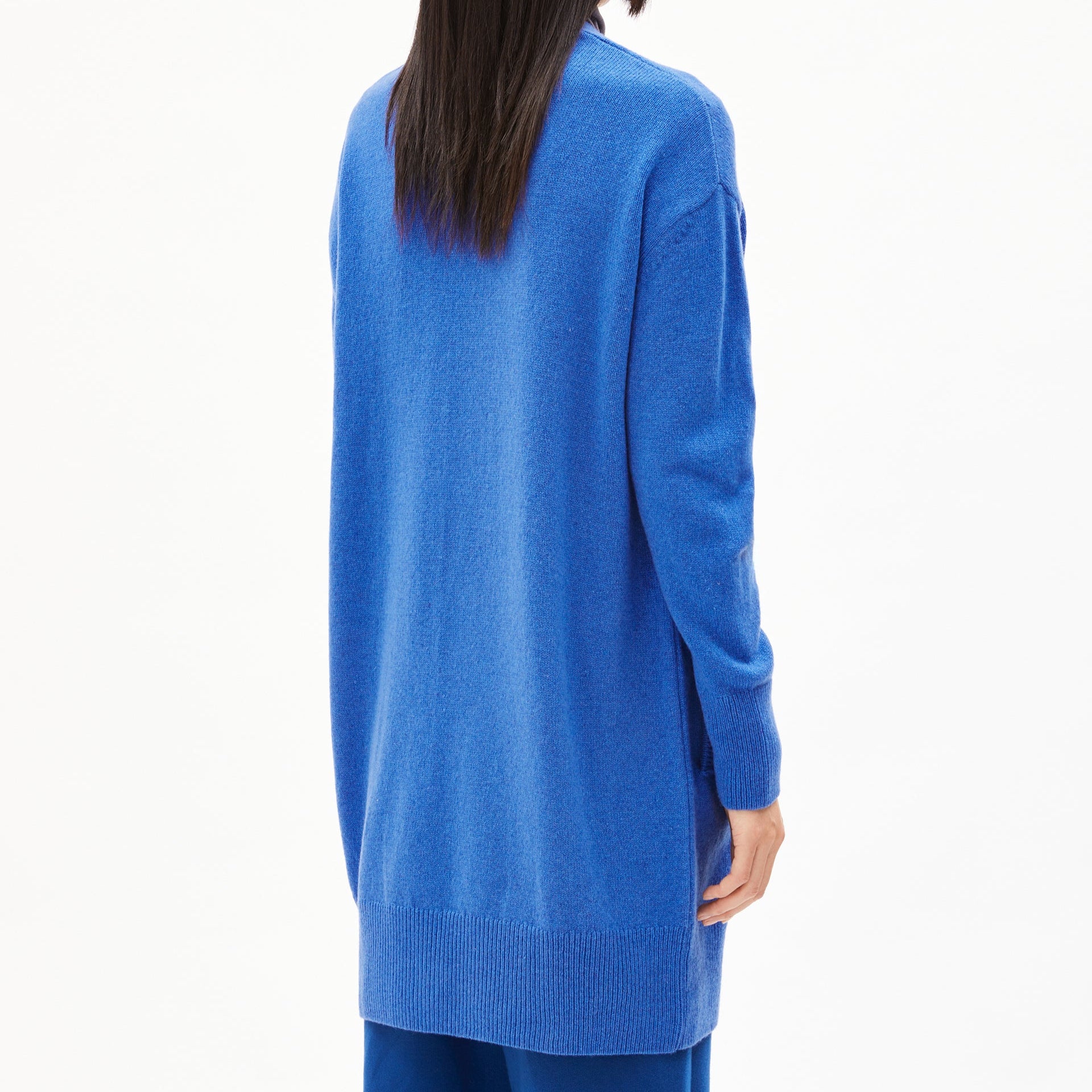 Person in einem langen, blauen Strickpullover, von hinten fotografiert, vor weißem Hintergrund.
