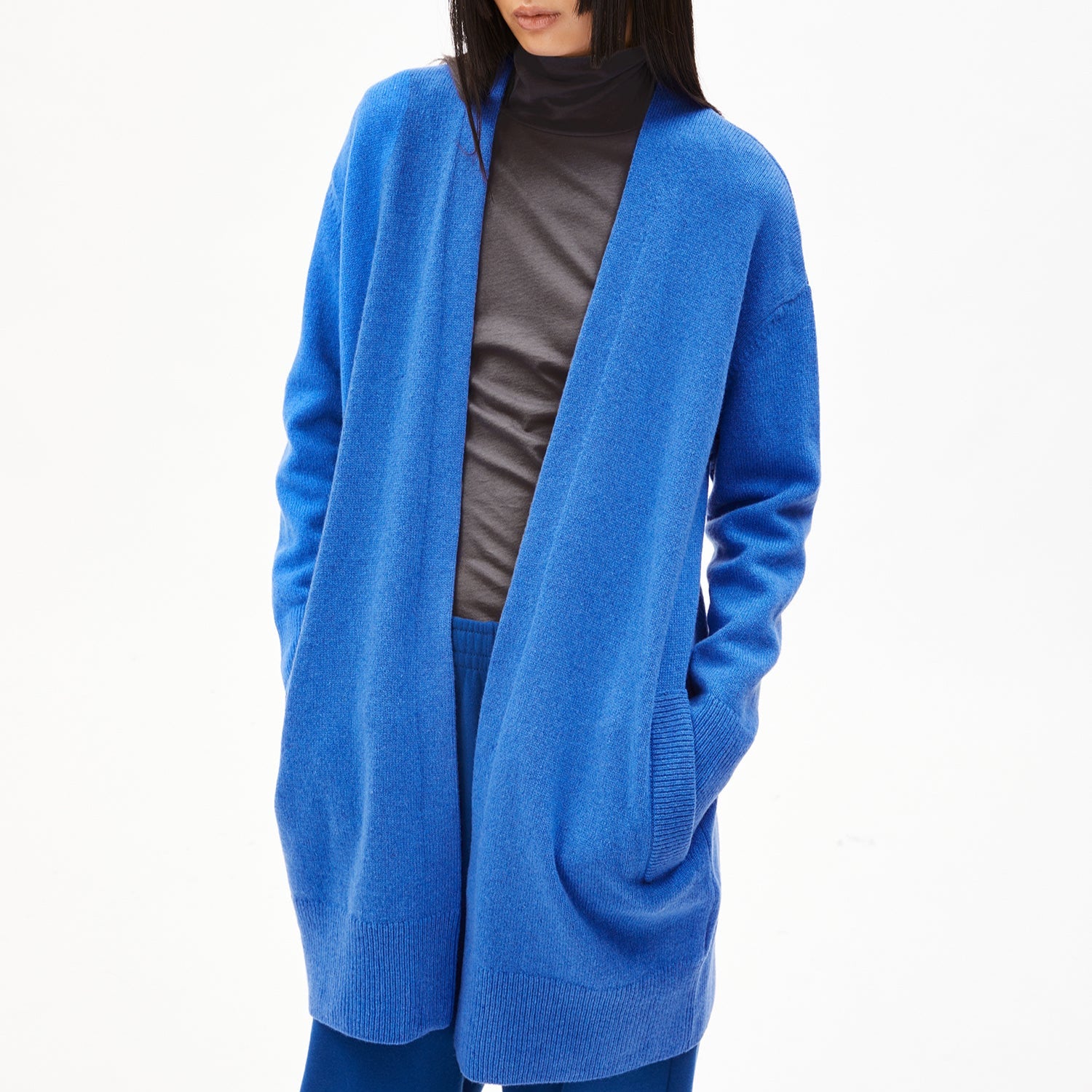 Person in blauem Cardigan und dunkelgrauem Rollkragenshirt, Hände in den Taschen, auf neutralem Hintergrund.