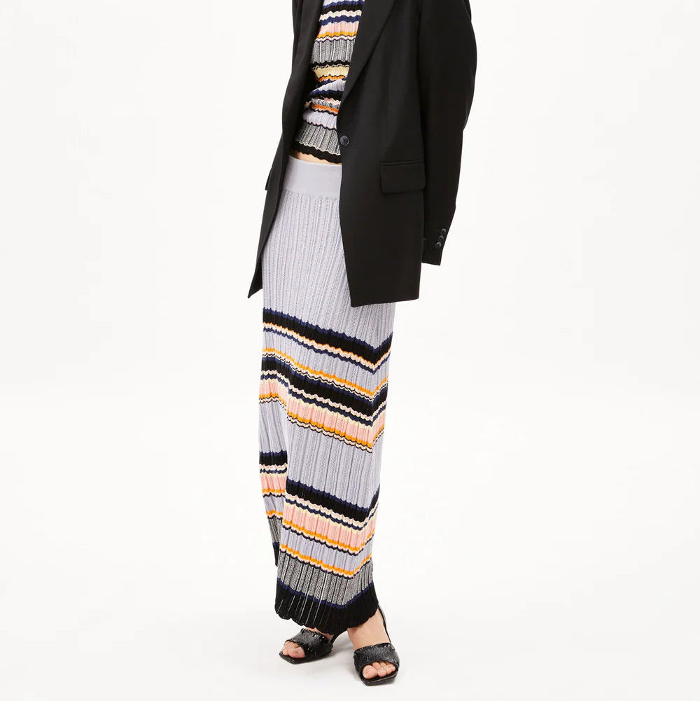 Eine Frau trägt einen schwarzen Blazer über einem gestreiften, gerippten Strickoberteil und einen passenden langen Rock in Grau-, Schwarz-, Gelb- und Weißtönen. Außerdem trägt sie schwarze, hochhackige Sandalen vor einem schlichten weißen Hintergrund.