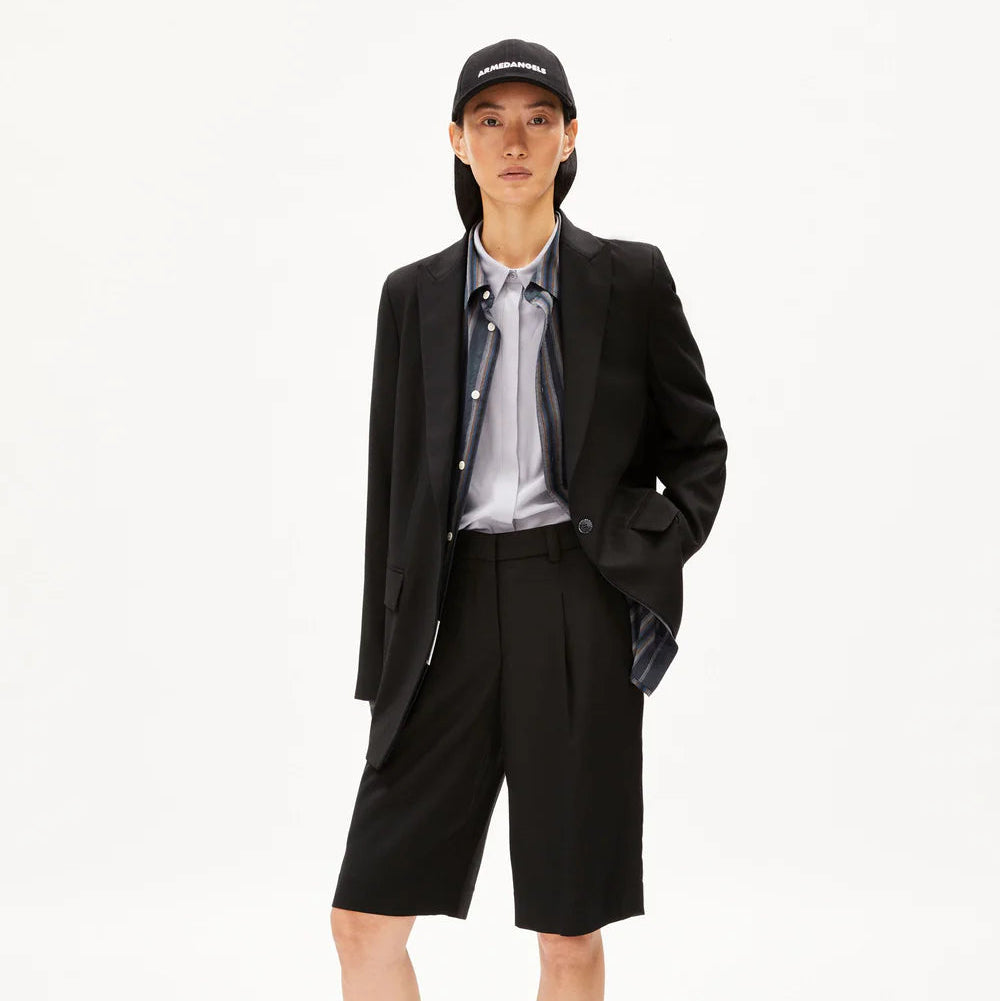 Eine Person, die einen schwarzen Blazer, ein hellblaues Hemd, lange schwarze Shorts und eine schwarze Baseballmütze trägt, steht vor einem schlichten weißen Hintergrund und hat eine Hand in der Tasche.