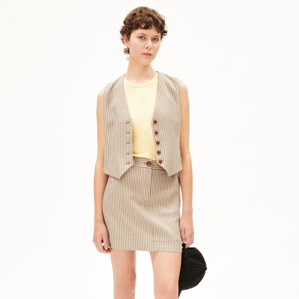 Eine Person mit kurzen, gelockten braunen Haaren trägt eine beige Weste mit Nadelstreifen und einen dazu passenden Minirock über einem gelben T-Shirt. Sie hält eine schwarze Tasche an ihrer Seite und steht vor einem schlichten weißen Hintergrund.