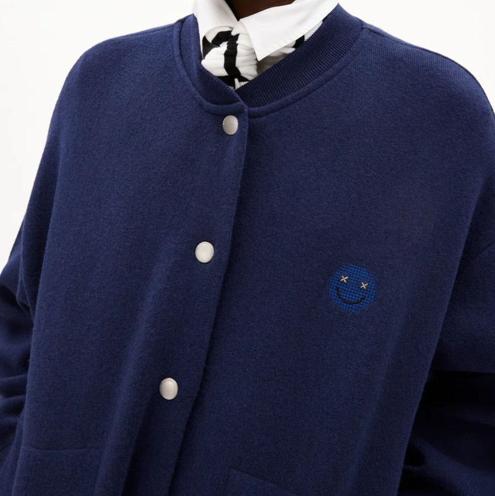 Eine Person, die eine schwarze Mütze, eine zugeknöpfte marineblaue Jacke mit einem kleinen Smiley-Logo und ein weißes Kragenhemd mit schwarzem Muster trägt, posiert vor einem schlichten weißen Hintergrund.