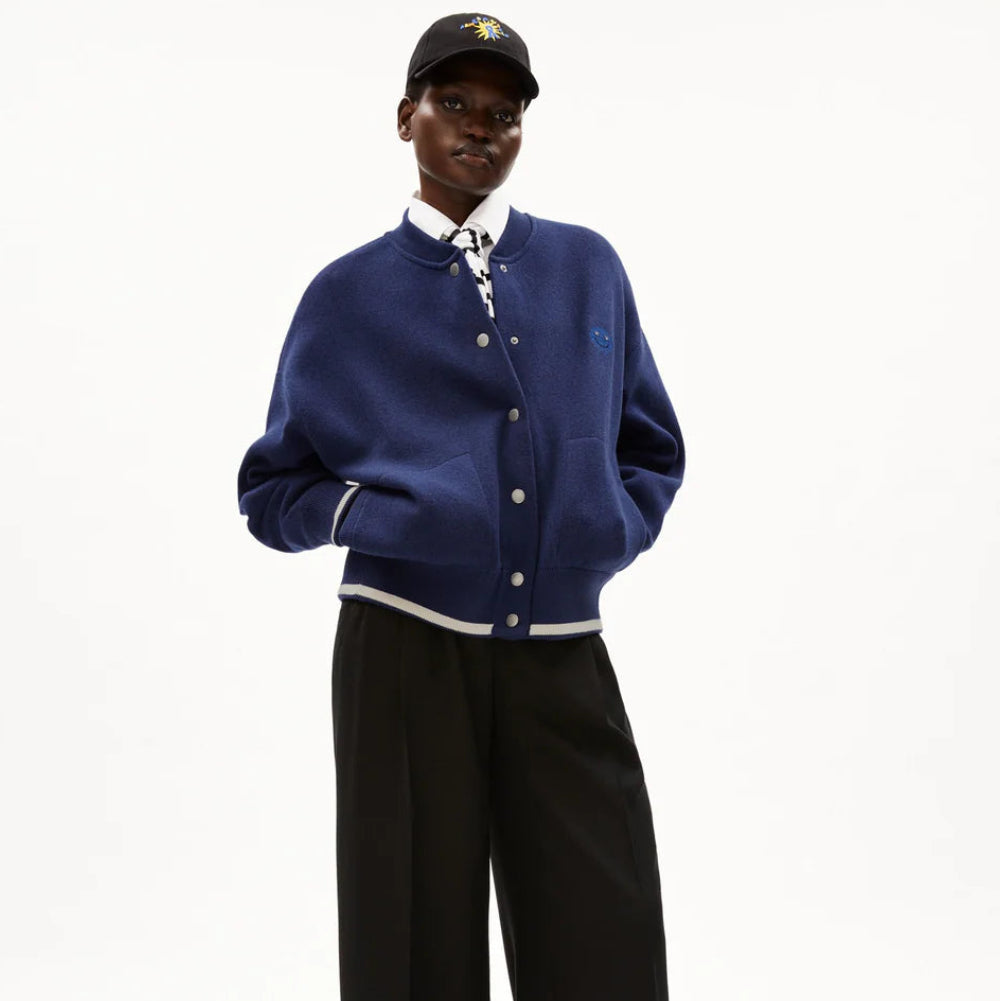 Eine Person, die eine marineblaue, zugeknöpfte Sportjacke, ein weißes Hemd, eine schwarze Hose mit weitem Bein, schwarze Schuhe und eine schwarze Baseballmütze trägt, steht mit den Händen in den Taschen vor einem schlichten weißen Hintergrund.