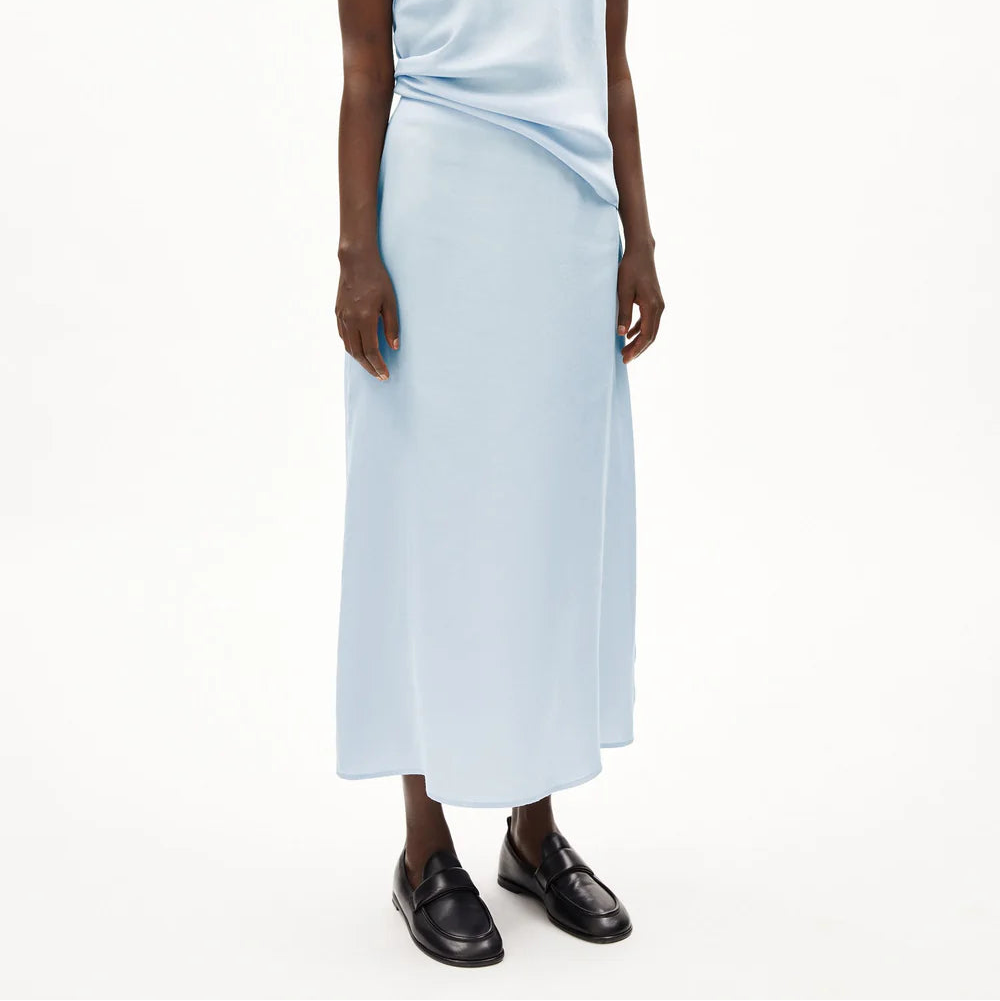 Eine Person trägt einen hellblauen, knöchellangen Rock und schwarze Halbschuhe, dazu ein passendes hellblaues Oberteil. Der Hintergrund ist einfarbig weiß. Der Kopf der Person ist auf dem Bild nicht zu sehen.