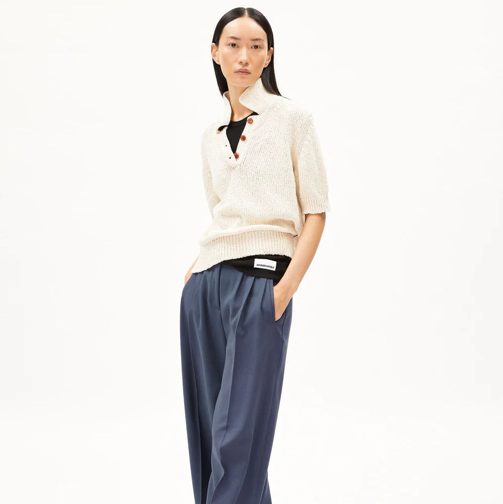 Eine Frau steht vor einem schlichten weißen Hintergrund, trägt ein kurzärmeliges cremefarbenes Strickpolo, eine weite blaue Hose und schwarze Schuhe, hat die Hände in den Taschen und einen neutralen Blick.