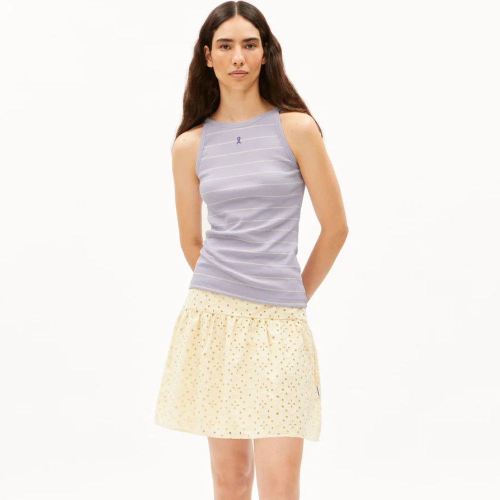 Eine Frau steht vor einem weißen Hintergrund und trägt ein hellviolettes ärmelloses Oberteil, einen gelben Rock mit kleinem Muster und transparente Jelly-Sandalen. Sie hat langes braunes Haar und einen neutralen Gesichtsausdruck.