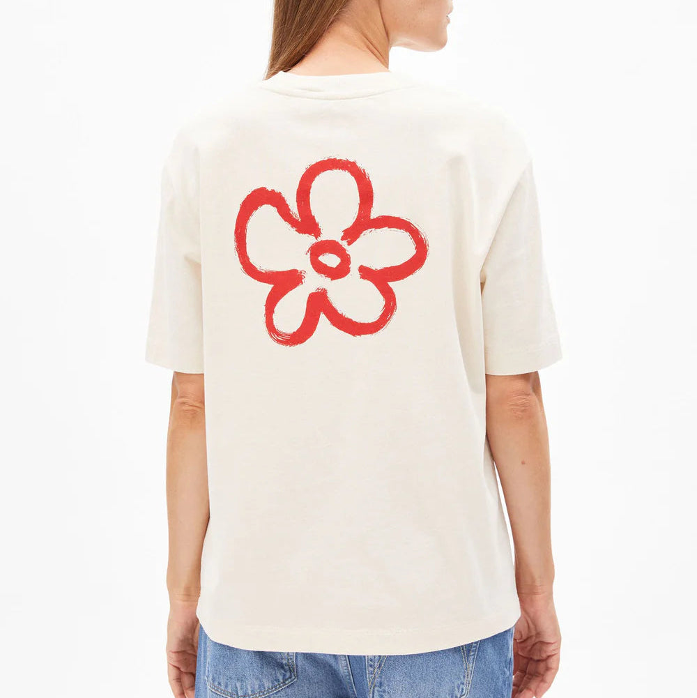 Eine Person mit glattem hellbraunem Haar trägt ein lockeres weißes T-Shirt mit einem großen, roten, handgezeichneten Blumenmuster auf dem Rücken, kombiniert mit einer lässigen Blue Jeans. Die Person blickt von der Kamera weg.