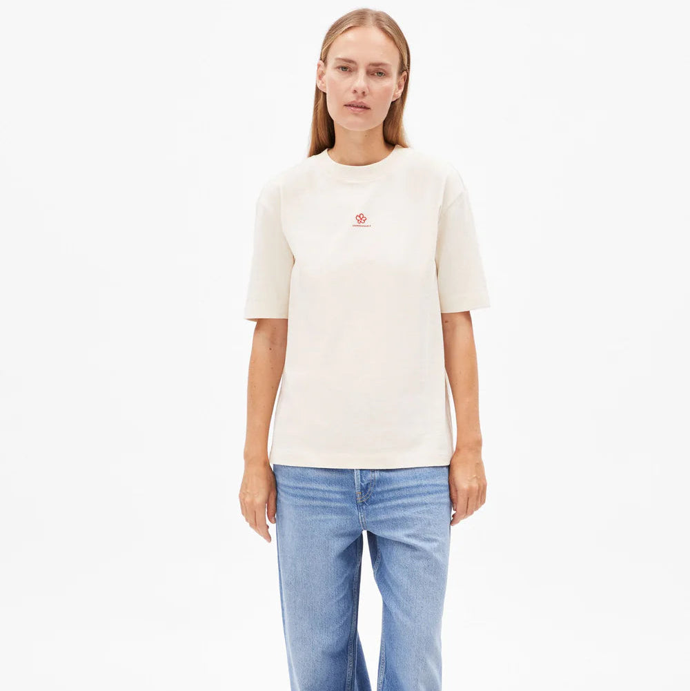 Eine Frau steht vor einem schlichten weißen Hintergrund und trägt ein cremefarbenes kurzärmeliges T-Shirt mit einem kleinen roten Logo, weite blaue Jeans und schwarze Flip-Flops. Sie hat glattes, hellbraunes Haar und einen neutralen Gesichtsausdruck.