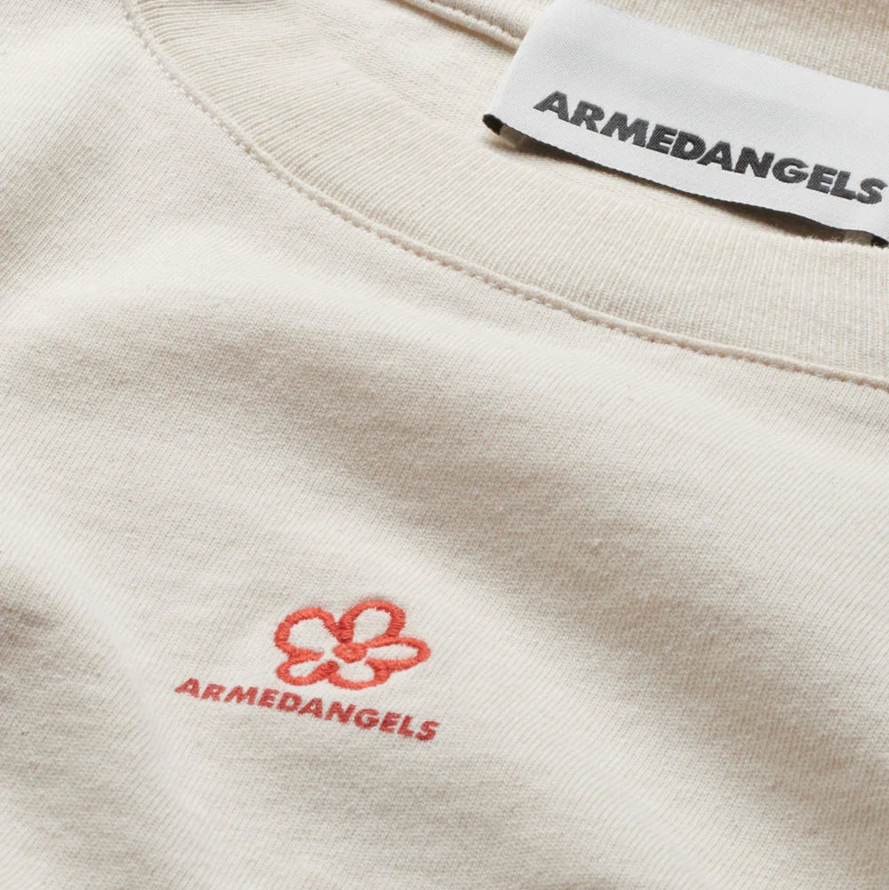 Nahaufnahme eines cremefarbenen ARMEDANGELS-Hemdes mit einer rot umrandeten Blume und dem gestickten Markennamen auf der Brust, wobei das ARMEDANGELS-Etikett am Halsausschnitt sichtbar ist.