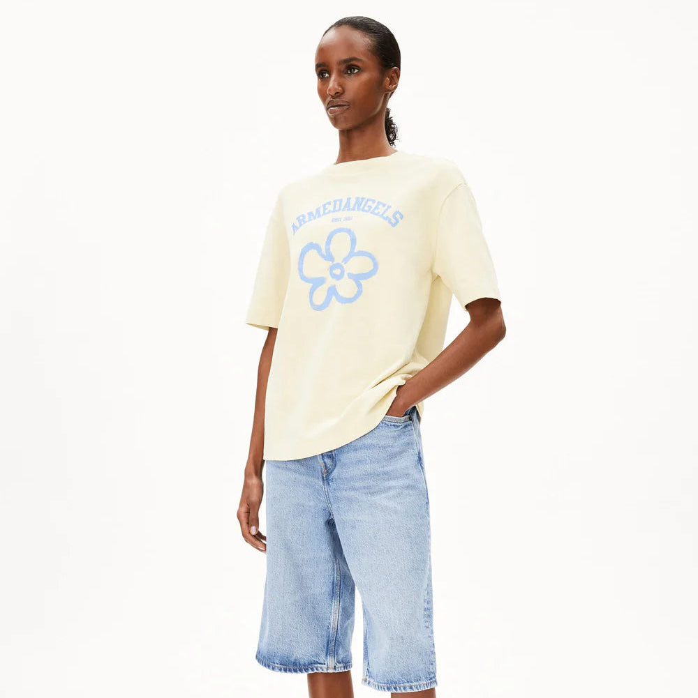 Eine Person, die ein hellgelbes T-Shirt mit einer blauen Blumengrafik und dem Wort ARMEDANGELS trägt, kombiniert mit knielangen hellblauen Jeans-Shorts, steht vor einem schlichten weißen Hintergrund.