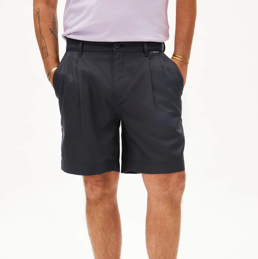 Eine Person, die ein hellviolettes Hemd trägt, das in eine hochtaillierte, dunkle, marineblaue Bundfaltenhose gesteckt ist und die Hände in den Taschen hat. Goldene Armbänder und Armtätowierungen sind sichtbar. Der Hintergrund ist weiß.